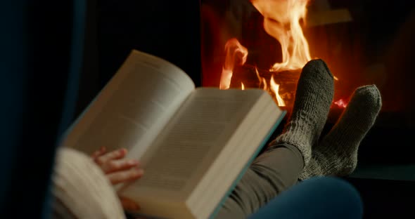 Woman Opens and Reads Book Relaxing at Fireplace in Cozy Room at Winter Night