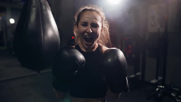 Portrait of Female Boxer Standing at Dark Gym Looking Intensely at the Camera and Screaming alt