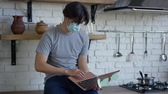 Young Asian Man in Covid Face Mask Texting Typing on Laptop Keyboard Sitting on Kitchen Countertop alt