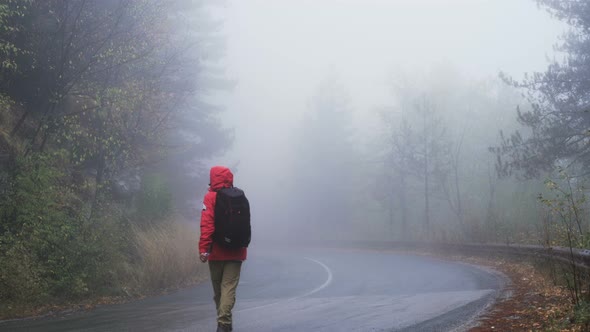 Male Tourist in Red Raincoat Walking on Foggy Wet Road alt