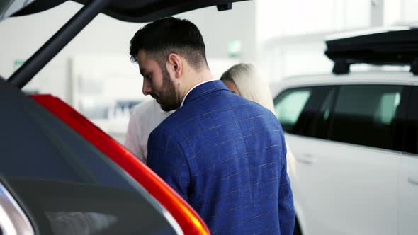 Salesman Showing Trunk From Inside to Customers in Automotive Dealership Showroom alt