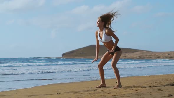 Girl in Sportswear Shorts and T-shirt Performs Jumps with Squats and Claps on the Beach Near the alt