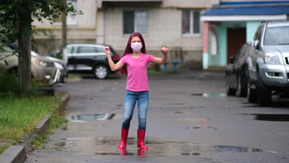 A little girl in a medical mask and rubber boots jumps through puddles in the courtyard