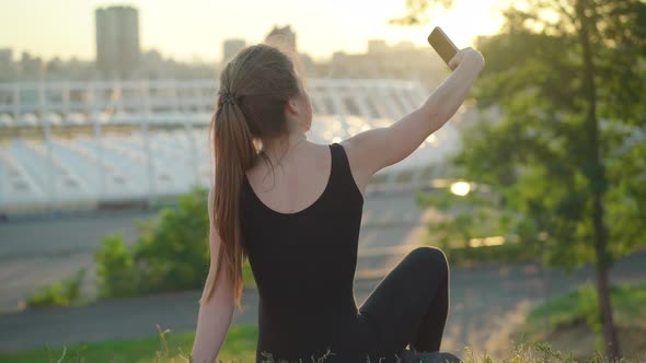 Back View of Young Caucasian Woman Sitting on Park Hill at Sunset and Taking Selfie. Carefree Slim alt