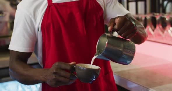 Midsection of african american barista pouring milk from jar into coffee cup in cafe alt