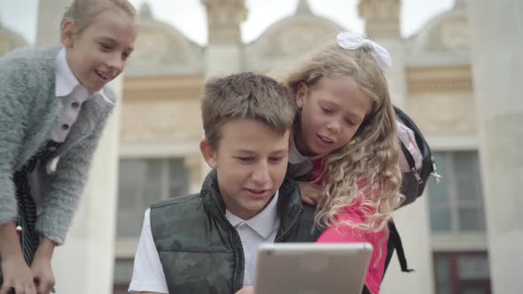 Three Cheerful Caucasian Schoolchildren Surfing Internet on Tablet During School Break. Portrait of alt