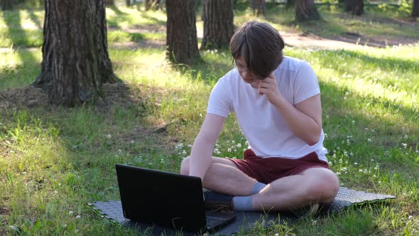 Young Man is Sitting on the Grass in Park with His Laptop and Talking on the Phone