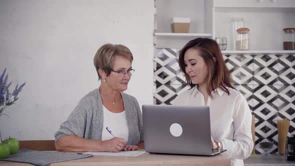 Adult Woman Using Laptop and Speaking with Senior Female on Kitchen