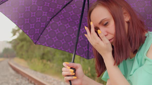 Young Woman with Umbrella Rubs Her Eyes Sitting on Railway in Cloudy and Windy Weather alt