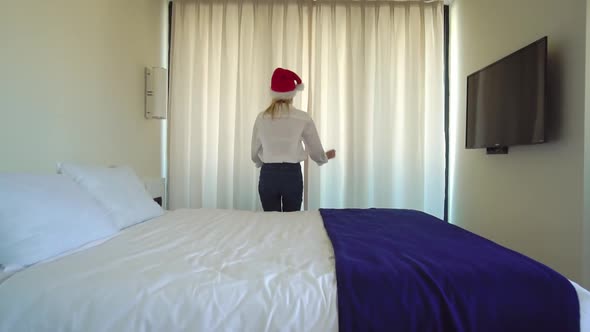 a woman in a Santa Claus hat gets out of bed and opens the window curtains. alt