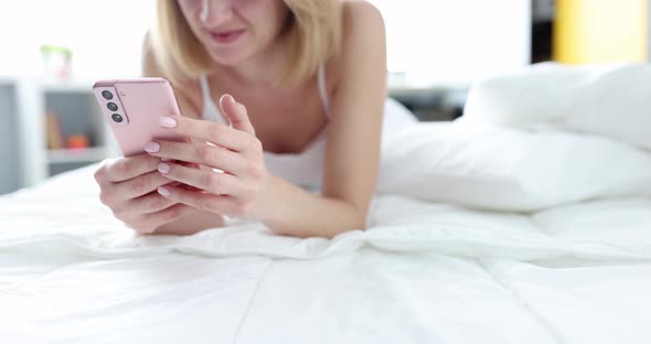 A Woman Lies on a Bed with a Smartphone in Her Hands alt
