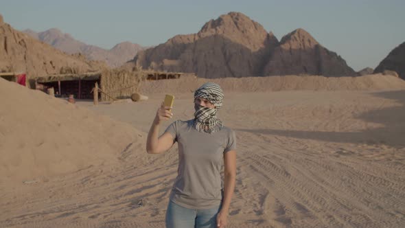 Tourist in Checkered Keffiyeh Trying to Find Mobile Network Using Cell Phone Standing in Desert alt