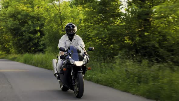 Man in Helmet Drives His Sport Black Motorcycle on the Summer Road Between Trees and Bushes During