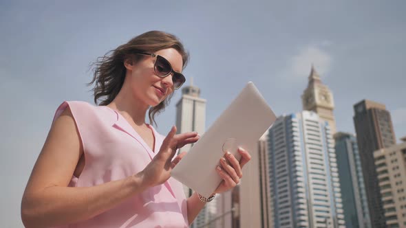 A Girl of European Appearance Works with a Tablet on the Background of Dubai Skyscrapers alt