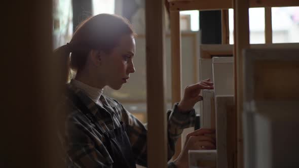 Focused Serious Young Female Artist Wearing Apron Choosing Best Canvas for Paintings in Her Dark alt