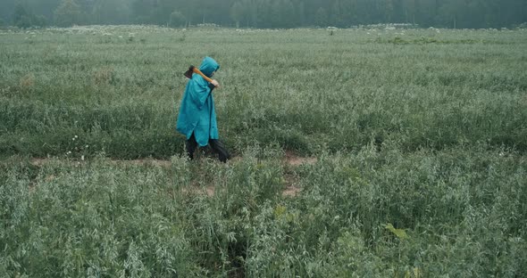 Woman in Blue Raincoat and Carrying an Axe is Walking Through the Field at Dusk