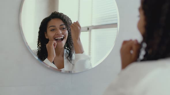 Young Positive African American Lady in Bathrobe Flossing Her Teeth Looking at Mirror at Bathroom in alt