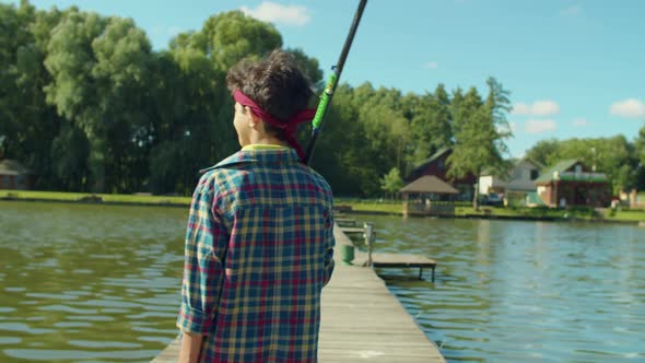 Rear View of Teenage Fisherman with Rod and Tackle Box Walking on Pier on Lake alt