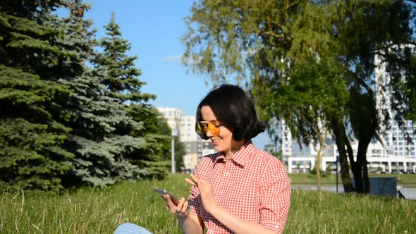 Young Attractive Woman in Summer Dress Sits on Green Grass with Mobile Phone alt