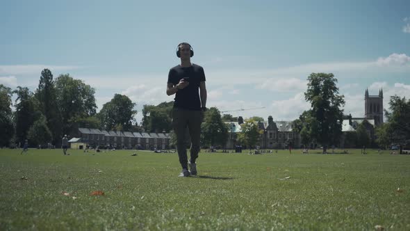 Young white man walking on the grass while checking his mobile phone in a park of Cambridge city, En alt