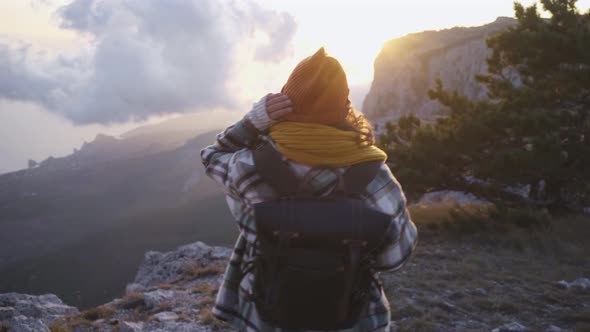 Happy Woman with Scarf and Hat Raises Hands on Steep Cliff