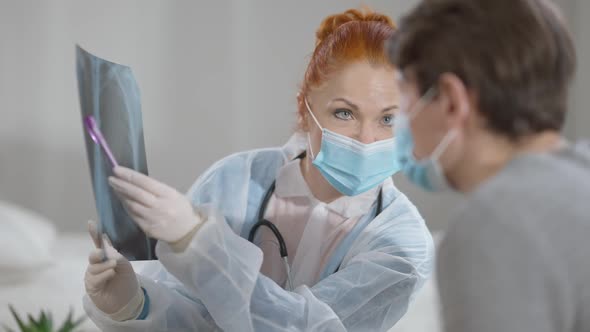 Portrait of Focused Caucasian Woman in Covid Face Mask Pointing at Lungs Xray Talking to Blurred Man alt