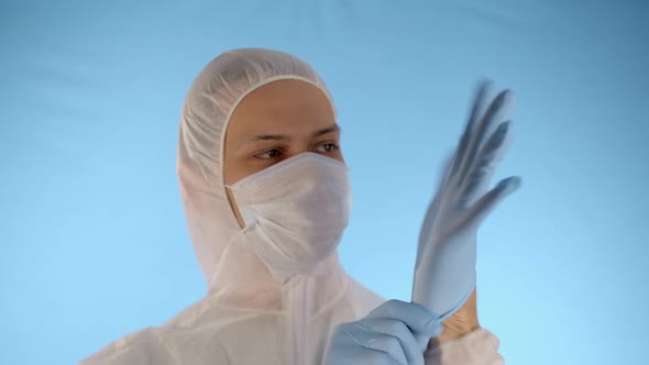 Man in Protective Suit and Medical Mask Puts Rubber Blue Gloves on His Hands