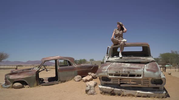 Young Woman in Safari Overalls Sits on the Rooftop of Abandoned Old Rusty Car alt
