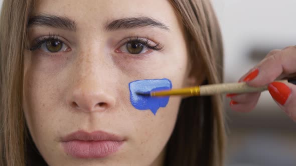 Close-up Portrait of Young Pleasant Woman Looking at Camera While Artist Painting with White on Blue alt