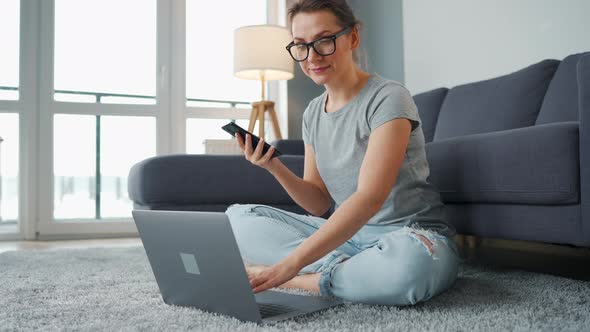 Casually Dressed Woman Sitting on Carpet with Laptop and Smartphone and Working in Cozy Room alt