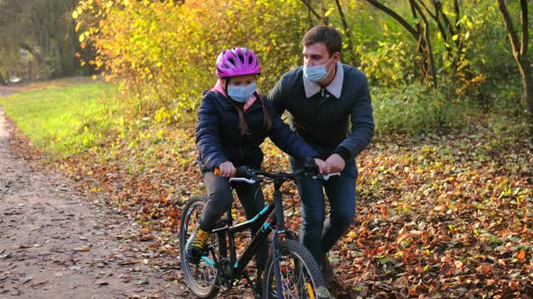Father and daughter in medical masks. Dad helps a girl learn to ride a bike