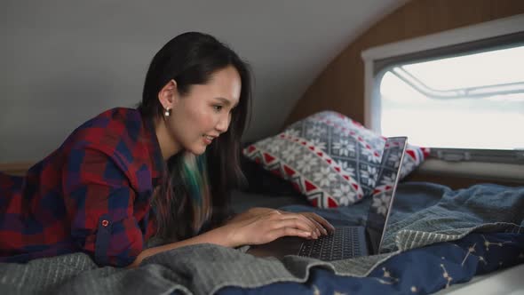 Asian Woman Freelancer Working at a Laptop While Lying in a Mobile Home Typing Text Remote Work in alt