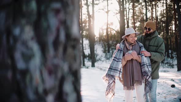 Man Covers a Woman with a Blanket in the Forest on a Winter Sunny Day