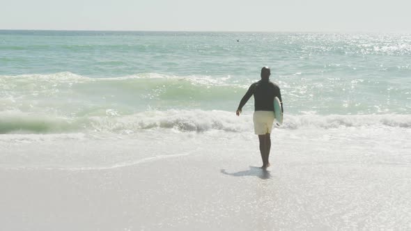 Senior african american man walking with surfboard on sunny beach alt