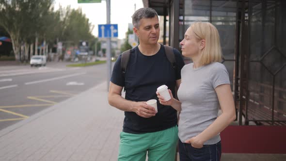 A Middleaged Man and Woman Drink Coffee From Paper Cups Stand at a Public Transport Stop Waiting for