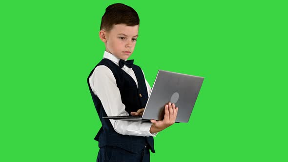 Concentrated Boy in a Bow Tie and Waistcoat Using Laptop Computer While Standing on a Green Screen alt
