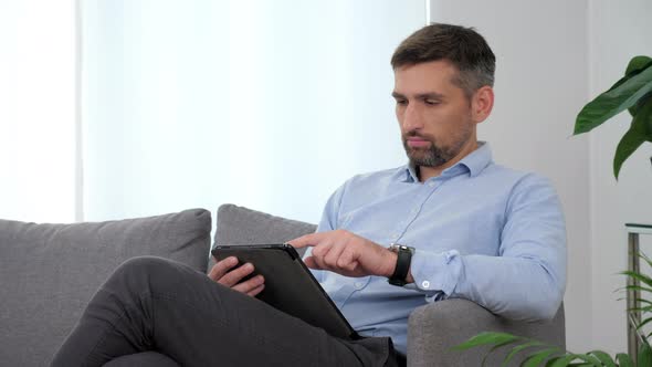 Businessman in Light Blue Shirt Sitting on the Couch Using Tablet Taping Screen alt