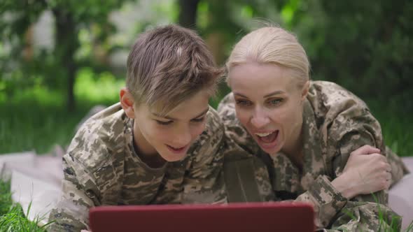 Carefree Military Woman and Teenage Boy Talking Laughing Surfing Internet on Laptop Lying on Sunny alt