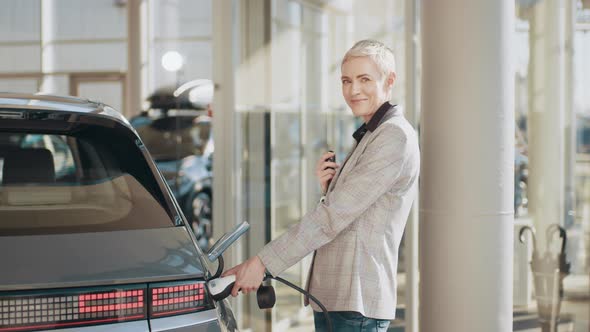 A Woman Near Her Electric Car Disconnects It From Charging in the Parking Lot alt