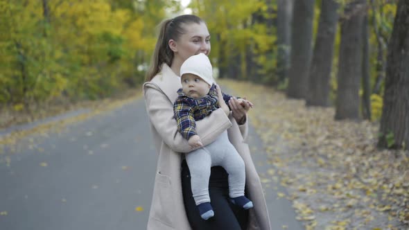 Mother Entertains Her Cute Baby Boy When Lulls Him on Hands During Walk in Park alt