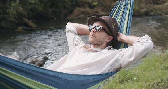 Close-Up View. Handsome Man in a Hat and Sunglasses Lying and Swinging in a Hammock on a Summer Day alt