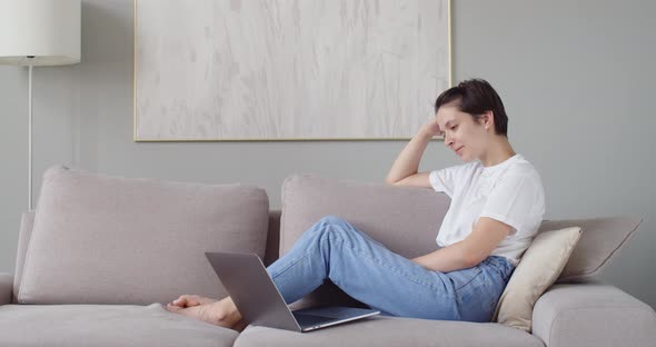 Attractive Girl Sits on Sofa Looking Funny Series on Laptop Spending Leisure Time