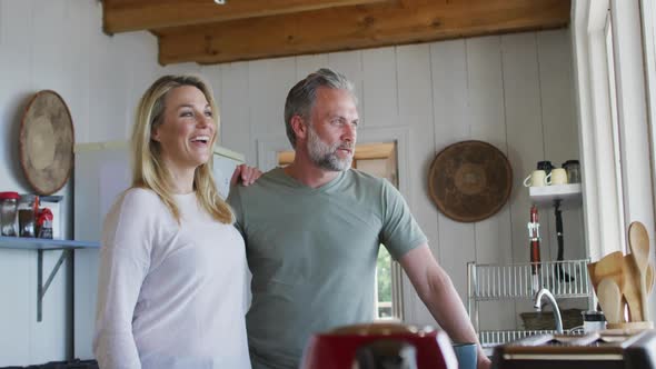 Happy caucasian mature couple drinking coffee and looking through the window in the kitchen alt