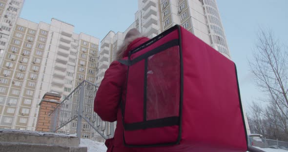 Follow Shot of Delivery Woman Wearing with Smartphone Red Uniform While Walking Along Buildings Down