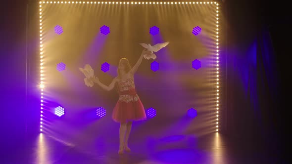 Young Attractive Woman Performs with Trained White Doves in Dark Studio Against the Backdrop of Blue alt