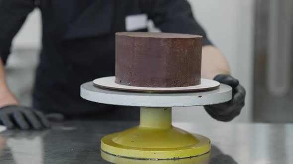 Blurred Caucasian Man in Coronavirus Face Mask Examining Round Chocolate Cake on Rotating Table alt