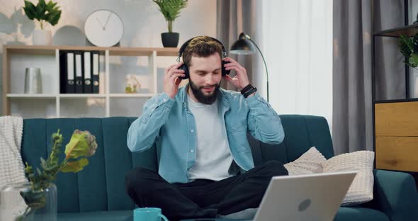 Man Sitting on Comfortable Sofa at Home and Enjoying Music from Phone Playlist Using Headphones alt