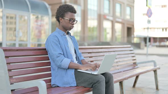 Coughing Young African Man Using Laptop While Sitting Outdoor on Bench alt