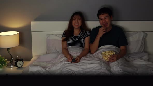 funny young couple watching TV and laughing on a bed at night