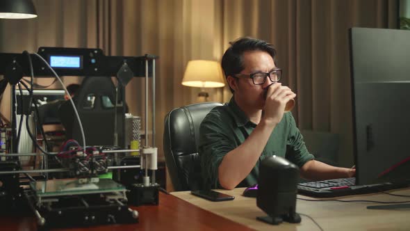 Asian Man Drinking Coffee While Works On Personal Computer And 3D Printer In Home Office alt